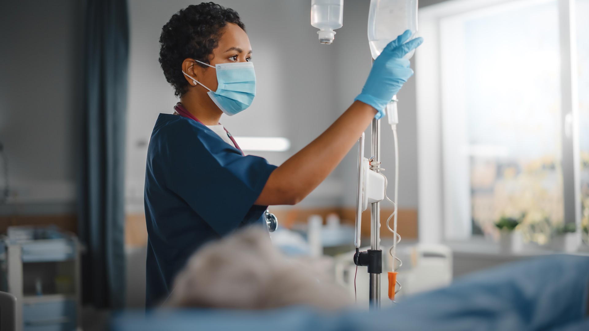 Hospital Ward: Professional Black Head Nurse Wearing Face Mask Does Checkup of Patient's Vitals, Checking Heart Rate Computer, Intravenous or Iv Fluids Drip Bag. Caring Nurse Monitors Person Recovery