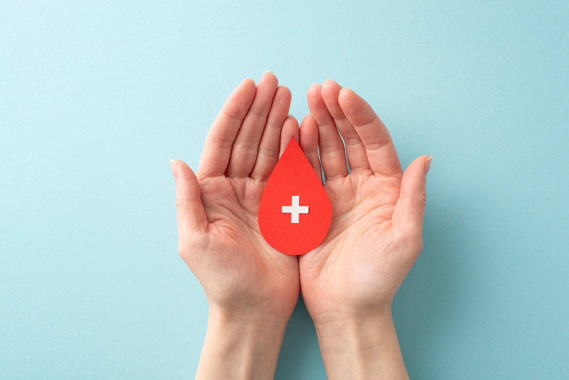 A first-person perspective top view photo of a female hands holding blood droplet with cross sign on palms on pastel blue background. It's the perfect visual for raising awareness about blood donation